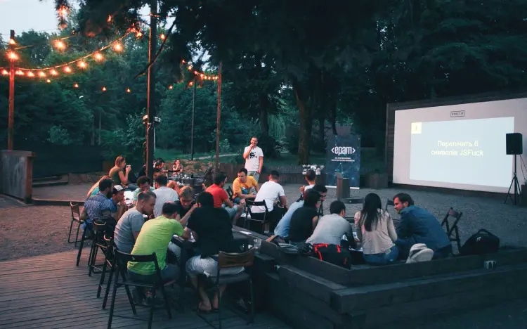 Outdoor event with people at tables listening to a presenter under string lights in the evening